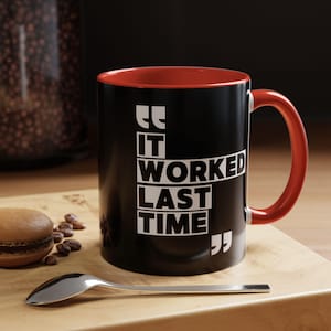 May include: Black coffee mug with a red interior and handle. The mug features the white text "IT WORKED LAST TIME" in a bold, modern font. The mug is on a wooden surface with a spoon, coffee beans, and a macaron.