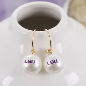 May include: A pair of gold-tone hoop earrings with white, pearl-like orbs. Each orb features the purple letters "LSU". The earrings are displayed against a white background.