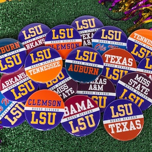May include: A collection of circular pin-back buttons featuring various college sports team names and logos. Each button displays the text House Divided along with team names like LSU, Auburn, Tennessee, Texas, Arkansas, and Alabama in bold colors.