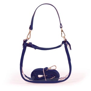 May include: A clear, transparent handbag with navy blue trim and a matching shoulder strap. The bag has a curved shape and gold-colored metal hardware. The shoulder strap is detachable and rolled up inside the bag.