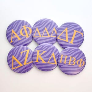 May include: Six circular pin-back buttons featuring a purple and lavender zebra stripe pattern. Each button displays gold Greek letters: Alpha Phi, Delta Delta Delta, Delta Gamma, Delta Zeta, Kappa Delta, and Pi Beta Phi, arranged in two rows of three.