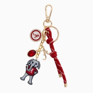 May include: Gold-tone keychain with a red and white cord, featuring charms. Charms include a red oval gem, a circular charm with a red "A" and rhinestones, and a cartoon elephant wearing a red shirt with an "A".