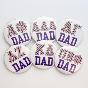 May include: Six circular white buttons featuring Greek letters in orange and purple diagonal stripes above the word DAD in bold purple block letters. The buttons display various Greek letter combinations including AΦ, ΔΔΔ, ΔΓ, ΔZ, KΔ, and ΠBΦ.