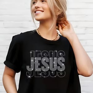 May include: Black t-shirt with the word "JESUS" in a rhinestone design. The word is stacked vertically with a sparkling effect. The shirt has short sleeves and a round neckline.