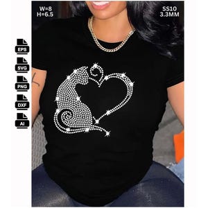 Op de afbeelding: Zwart t-shirt met een strass ontwerp van een kat die een hart vormt. De kat en het hart zijn gemaakt van kleine, glinsterende strass steentjes. Het shirt heeft korte mouwen en een ronde hals. Afmetingen ontwerp: B=20.3 cm, H=16.5 cm.