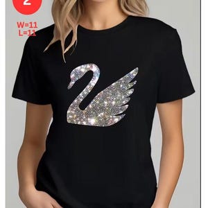 May include: Black t-shirt featuring a sparkling swan design. The swan is made of iridescent rhinestones. The text "2" is in a red circle in the upper left corner. The text "W=11 L=11" is below the red circle.