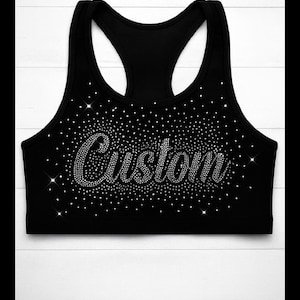 May include: Black athletic crop top with a racerback design. The word "Custom" is spelled out in sparkling rhinestones across the front, surrounded by a scattering of smaller rhinestones. The top is set against a white wooden background.