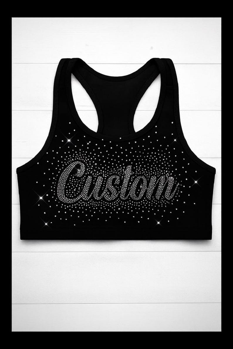 May include: Black athletic crop top with a racerback design. The word "Custom" is spelled out in sparkling rhinestones across the front, surrounded by a scattering of smaller rhinestones. The top is set against a white wooden background.