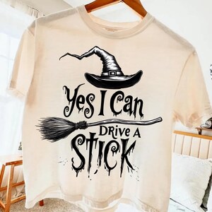 Peut inclure: T-shirt crème avec un chapeau de sorcière noir et un motif de balai. Le texte "Yes I Can Drive A Stick" est imprimé sous le chapeau et le balai. Le t-shirt a des manches courtes et un col rond.