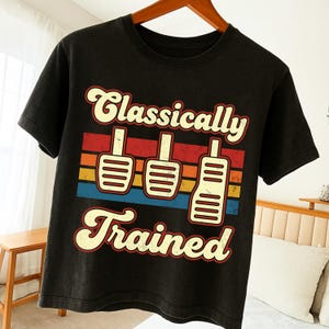 Może przedstawiać: Czarny t-shirt z napisem "Classically Trained" w stylu retro. Projekt zawiera trzy białe pedały samochodowe na paskowanym tle w kolorach czerwonym, pomarańczowym, żółtym i niebieskim.