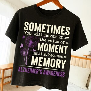 Peut inclure: T-shirt noir avec l'inscription "SOMETIMES You will never know the value of a MOMENT until it becomes a MEMORY ALZHEIMER'S AWARENESS". Le motif comprend des pissenlits et des papillons violets.