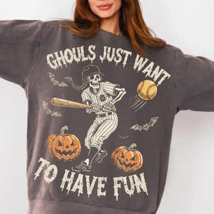 Peut inclure: Sweat-shirt gris foncé avec un motif sur le thème d'Halloween. Le motif représente un joueur de baseball squelette frappant une batte, une balle de baseball, des chauves-souris et des citrouilles. Le texte dit "Ghouls Just Want To Have Fun."