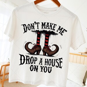 Può includere: T-shirt bianca con grafica a tema Halloween. Il design presenta un paio di scarpe da strega marroni con punte ricciolute, gambe a righe rosse e nere e la scritta "DON'T MAKE ME DROP A HOUSE ON YOU" in un font gotico.