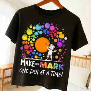 Peut inclure: T-shirt noir avec un graphisme coloré. Le motif comprend un grand cercle orange, un petit personnage de dessin animé tenant un pinceau et de nombreux points multicolores. Le texte "MAKE YOUR MARK ONE DOT AT A TIME!" est également inclus.