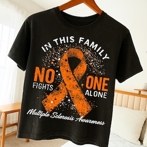 Peut inclure: T-shirt noir avec un ruban orange et le texte "IN THIS FAMILY NO ONE FIGHTS ALONE" et "Multiple Sclerosis Awareness" en blanc. Le t-shirt est suspendu à un cintre en bois.