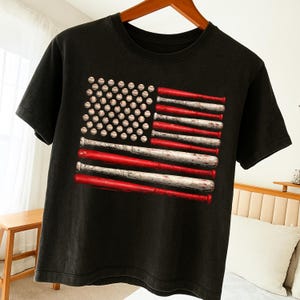 Peut inclure: T-shirt noir avec un motif du drapeau américain composé de balles et de battes de baseball. Les étoiles sont des balles de baseball, et les rayures sont des battes rouges et blanches. Le t-shirt a un aspect vintage et usé.