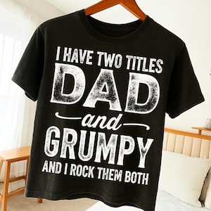 Peut inclure: T-shirt noir avec texte blanc : "I HAVE TWO TITLES DAD and GRUMPY AND I ROCK THEM BOTH". Le t-shirt est suspendu à un cintre en bois. Le t-shirt est de style col rond.