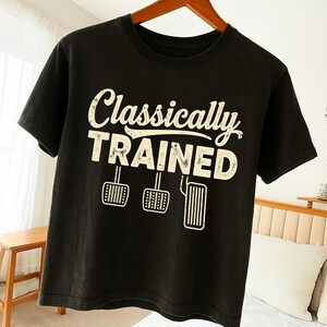 Może przedstawiać: Czarny t-shirt z napisem "Classically TRAINED" w postarzanym, złamanym białym kolorze. Poniżej tekstu znajdują się trzy ilustracje pedałów. Koszulka wisi na drewnianym wieszaku.