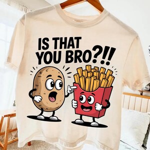 May include: Cream-colored t-shirt featuring a cartoon potato and a box of fries. The potato has a surprised expression, and the fries are pointing at it. The text "IS THAT YOU BRO?!" is printed above the characters.