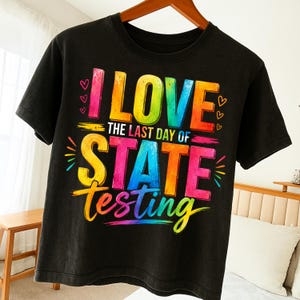 Peut inclure: T-shirt noir avec l'inscription colorée "I LOVE THE LAST DAY OF STATE TESTING". Le texte est multicolore avec des cœurs et des éclats d'étoiles. Le t-shirt est suspendu à un cintre en bois.