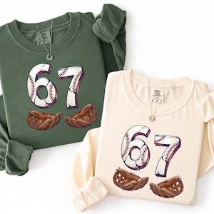 May include: Two long-sleeved sweatshirts, one green and one cream, each with the number 67 designed as a baseball. Below each number is a baseball glove. Both shirts have a necklace.