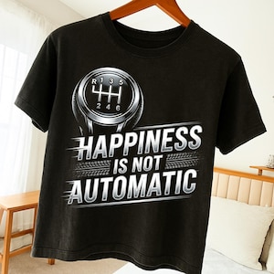 May include: Black t-shirt featuring a silver gear shift graphic with the text "HAPPINESS IS NOT AUTOMATIC". The design includes a tire track element. The shirt is displayed on a wooden hanger.