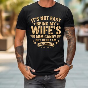 Peut inclure: T-shirt noir avec l'inscription dorée "IT'S NOT EASY BEING MY WIFE'S ARM CANDY BUT HERE I AM NAILING IT". Le texte est mis en valeur par des étoiles et des ailes, pour un design audacieux.