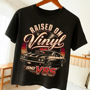 Puede incluir: Camiseta negra con un diseño gráfico de un coche clásico y el texto "RAISED ON Vinyl AND V8S". El diseño incluye un disco de vinilo y un fondo tipo puesta de sol. La camiseta está colgada en una percha de madera.