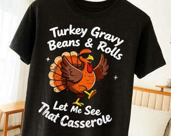 May include: Black t-shirt featuring a cartoon turkey wearing sunglasses, with the text "Turkey Gravy Beans & Rolls Let Me See That Casserole". The turkey is orange, brown, and yellow, with its wings spread. A fun, festive design.