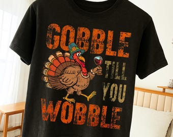 May include: Black t-shirt with a distressed orange graphic that reads "GOBBLE TILL YOU WOBBLE." The graphic features a cartoon turkey wearing a pilgrim hat and holding a glass of wine. The shirt is hanging on a wooden hanger.