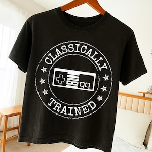 May include: Black t-shirt with a white graphic of a retro video game controller inside a circular design. The text "CLASSICALLY TRAINED" is printed around the controller. The shirt is hanging on a wooden hanger.
