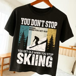 May include: Black t-shirt with a skiing graphic. The design includes the text "YOU DON'T STOP SKIING WHEN YOU GET OLD, YOU GET OLD WHEN YOU STOP SKIING" in white. The graphic features a skier on a slope with trees.