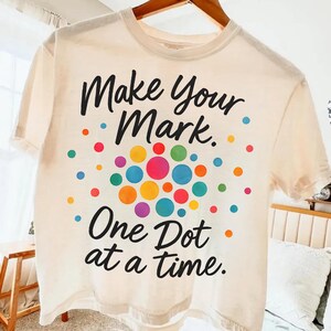 Peut inclure: T-shirt crème avec le texte "Make Your Mark. One Dot at a Time." en écriture noire. Le t-shirt présente un motif de points colorés dans des tons de bleu, vert, jaune, orange, rose et rouge.