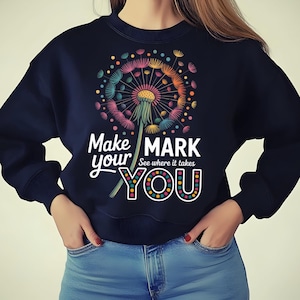 Peut inclure: Sweat-shirt bleu marine avec un motif de pissenlit coloré et l'inscription "Make your mark, see where it takes you". Le pissenlit présente des accents roses, oranges, jaunes et bleus. Le mot "YOU" est orné de points.