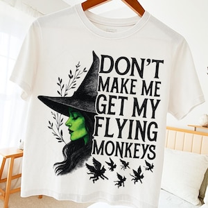 May include: White t-shirt featuring a graphic of a green-skinned witch wearing a black hat. The text "DON'T MAKE ME GET MY FLYING MONKEYS" is printed in black. The design includes black flying monkey silhouettes and leafy sprigs.