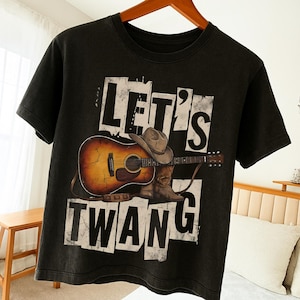 Puede incluir: Camiseta negra con un gráfico desgastado que dice "LET'S TWANG". El diseño incluye una guitarra acústica marrón, un sombrero de vaquero y botas. La camiseta está colgada en una percha de madera.