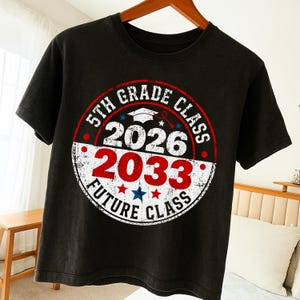 Peut inclure: T-shirt noir avec un motif circulaire. Le motif indique "5th Grade Class 2026 2033 Future Class" en rouge, blanc et bleu. Une toque de diplômé et des étoiles sont également incluses. Le t-shirt est suspendu à un cintre en bois.