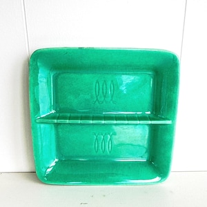 May include: A rectangular, emerald green ceramic soap dish. The dish has a raised divider and decorative swirl designs. The glossy finish reflects light, highlighting the vibrant color and simple design.