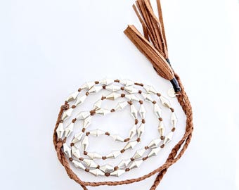 May include: A long necklace featuring silver-colored geometric beads strung on a brown cord. The necklace has a braided cord and a brown leather tassel at the end. The jewelry is displayed on a white background.