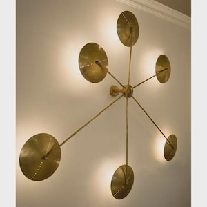 May include: A brass wall light with six circular, perforated shades. The fixture features a central hub with radiating arms, each ending in a disc-shaped shade. The shades are lit from behind, creating a warm glow, ideal for a living room.