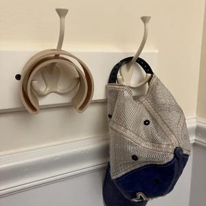 May include: A white wall-mounted hat rack with two beige hooks. One hook holds a beige and navy blue baseball cap. The other hook holds a beige hat holder.