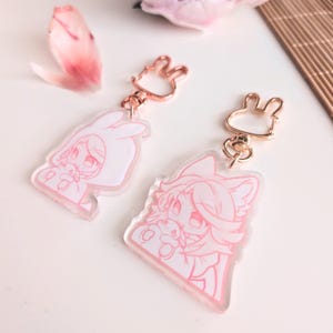 Könnte beinhalten: Zwei rosa Acryl-Schlüsselanhänger mit Cartoon-Charakter-Designs. Ein Schlüsselanhänger zeigt einen Charakter mit Hasenohren, der andere einen Charakter mit Fuchsohren. Beide haben goldfarbene, hasenförmige Verschlüsse. Die Schlüsselanhänger liegen auf einer weißen Oberfläche.