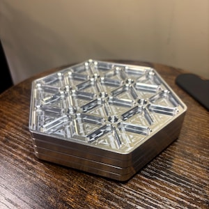 May include: A silver, hexagonal metal box with a geometric design on the top surface. The box is made of multiple layers and sits on a dark wooden surface. The design features repeating triangular and circular patterns.