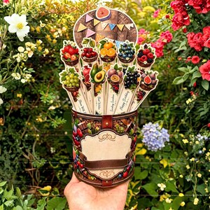 May include: A cylindrical container with a brown woven pattern and a central blank label, holding multiple vertical paper sticks topped with illustrations of various fruits in baskets, including strawberries, cherries, oranges, and grapes.