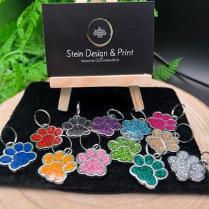 Puede incluir: Llaveros con forma de huella de pata con purpurina en varios colores: azul, rojo, naranja, rosa, verde y plateado. Un pequeño letrero con el texto "Stein Design & Print" se encuentra en el fondo.