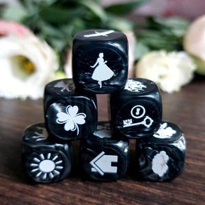 May include: A stack of seven black dice with white symbols, including a bird, a woman in a dress, a four-leaf clover, a key, a sun, a house, and a cloud. The dice are arranged on a dark wooden surface, with flowers in the background.