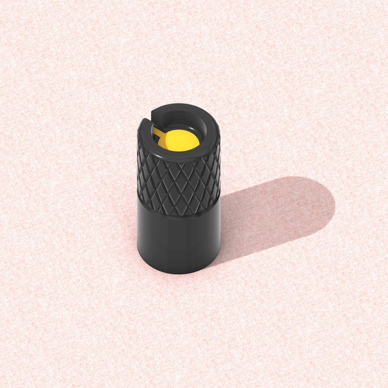 以下が含まれることがあります： 上部にダイヤモンド模様、内部に黄色のインサートが付いた黒いタイヤバルブステムキャップ。 キャップは円筒形で、ピンク色のテクスチャ加工された表面に置かれ、影を落としています。