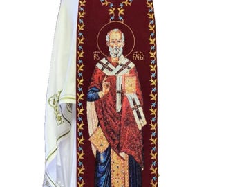 Pode incluir: Uma estola litúrgica com um painel central bordô com uma figura religiosa bordada. A estola é adornada com elementos decorativos dourados e azuis, incluindo padrões florais e borlas. A vestimenta branca tem acabamentos dourados.