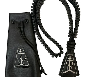 Puede incluir: Una bolsa negra con cordón, superficie texturizada y un emblema de cruz bordado en blanco. Se acompaña de una correa larga de cuentas negras con un parche triangular a juego que presenta un bordado blanco de una cruz y pequeñas cruces.