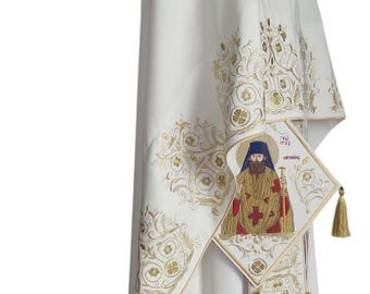 May include: White liturgical vestment with gold embroidery. The ornate design features floral patterns and a religious icon. The vestment includes a matching stole with tassels. The overall design is elegant and detailed.
